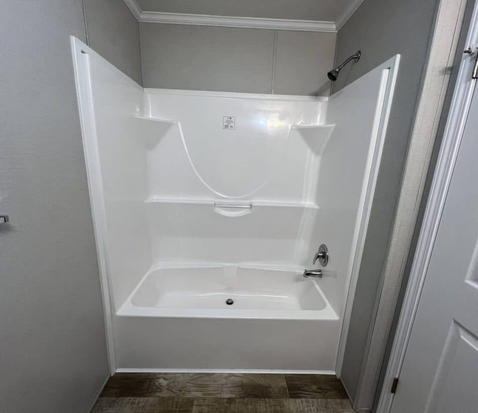 A white bathtub and shower combo with built-in shelves in a small, clean bathroom, surrounded by light gray walls and wood-style flooring. A towel bar is mounted on the left wall, and a white door is on the right.