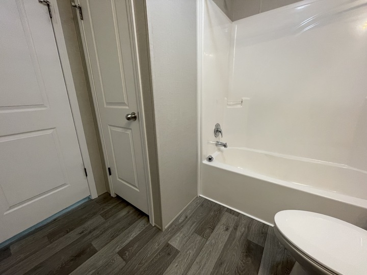 A clean bathroom with wood-look flooring, white walls, a white bathtub, a toilet, and two closed doors. The space appears modern and well-lit.