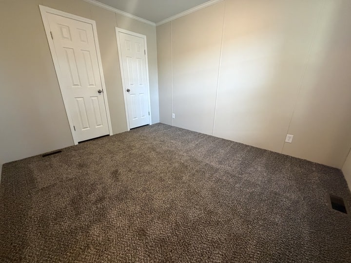 Empty room with beige walls, brown carpet, and two closed white doors. There are electrical outlets and a vent on the floor near the walls. The room is well-lit with natural light.