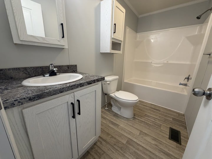 A modern bathroom with light wood cabinets, a marble-look countertop with a sink, a toilet, and a white bathtub with shelves. The floor has wood-look tiles and the walls are light gray.