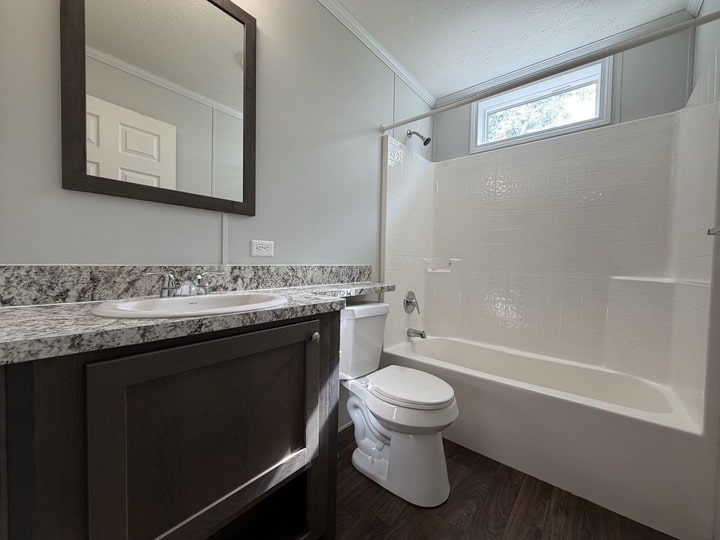 A modern bathroom with a granite countertop, sink, dark wooden vanity, white toilet, and a white bathtub-shower combo. There is a large mirror above the sink and a small window above the tub.