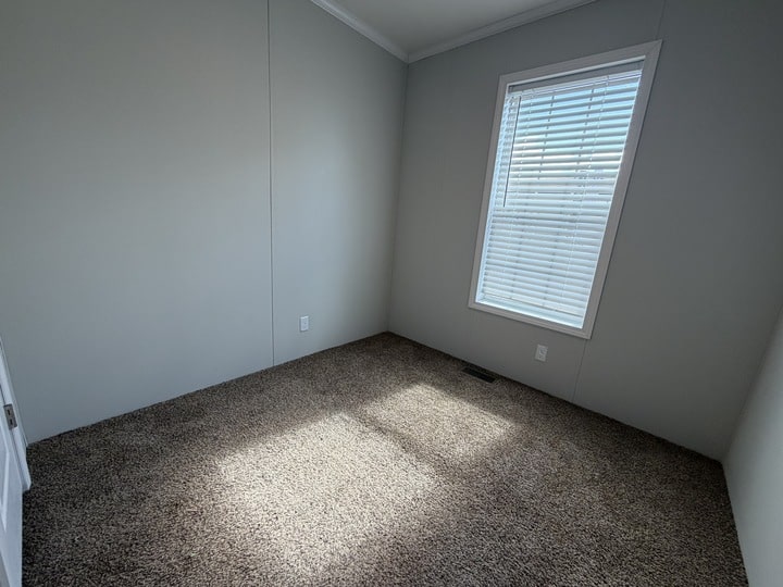 Small empty room with gray walls, a window with white blinds letting in sunlight, and brown carpeted flooring. The room appears clean and minimal, with natural light creating a bright atmosphere.