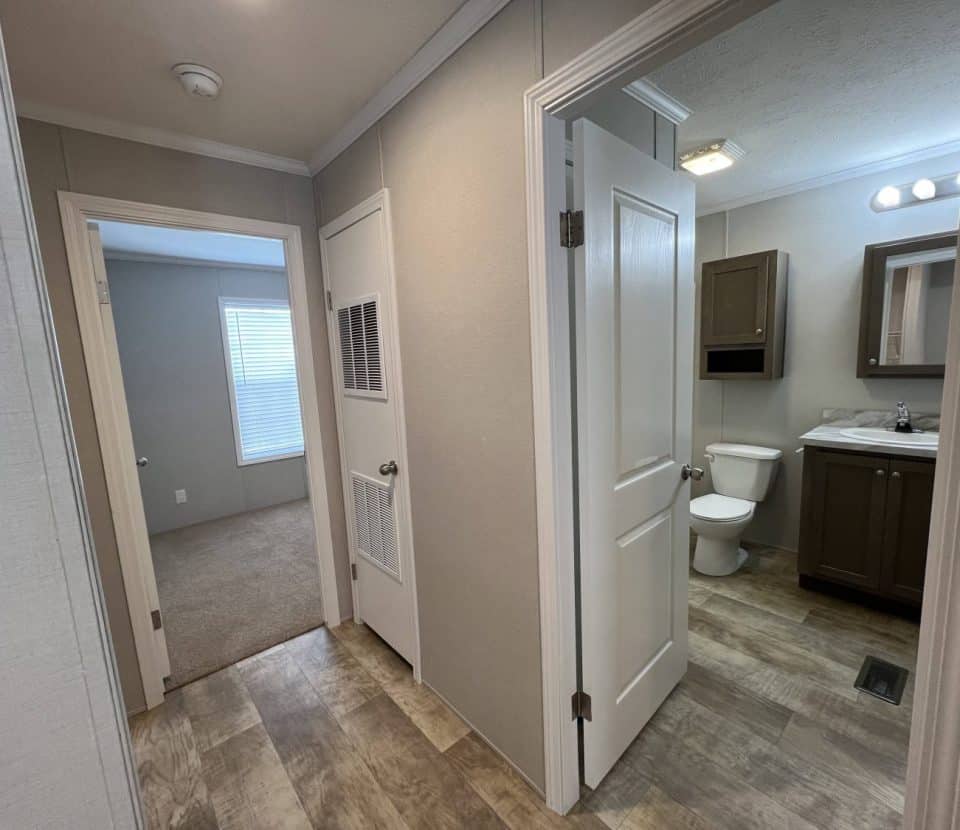 A hallway with light wood flooring leads to a carpeted bedroom on the left and a bathroom with brown cabinets, a toilet, and a sink with a mirror on the right. Walls are beige with white trim.