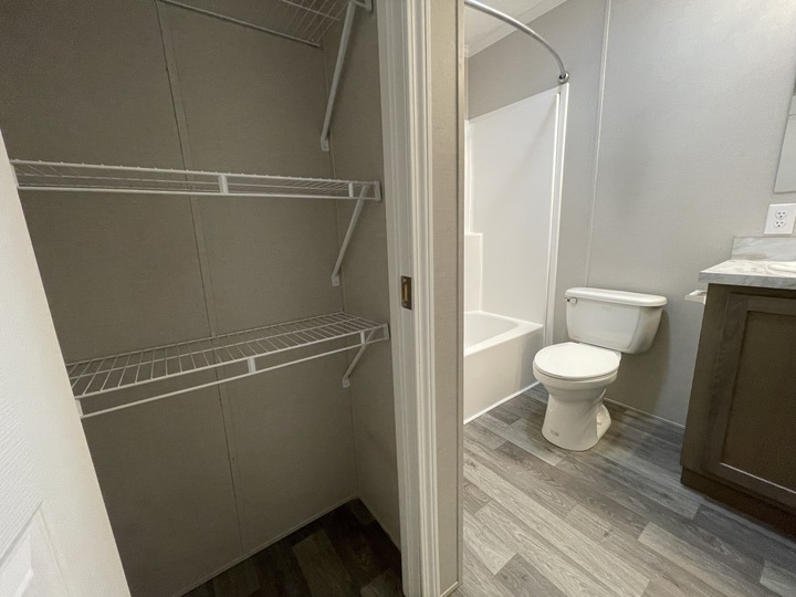 A small bathroom with gray flooring, a white toilet, a bathtub with a shower curtain rod, a vanity with a sink, and an open closet with several white wire shelves.