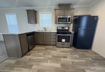 Modern kitchen with wood cabinets, marble countertops, stainless steel dishwasher, oven, and microwave, and a black refrigerator. The floor is wood-patterned, and two windows let in natural light.