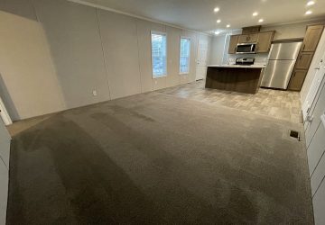Open-plan living area with carpeted floor leading to a kitchen featuring wood cabinets, stainless steel appliances, and tile flooring. Two windows let in natural light; recessed ceiling lights provide illumination.