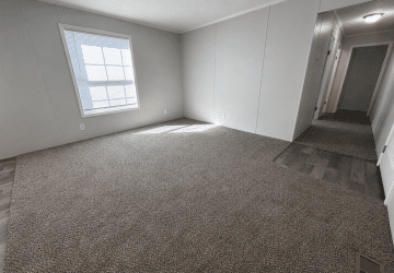 Empty room with light gray walls, a large window letting in sunlight, carpeted flooring, and a hallway with wood-style flooring leading to other rooms. The space appears clean and unfurnished.