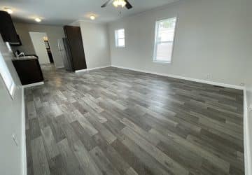 Empty room with light-colored walls, dark trim, wood-look vinyl flooring, two large windows letting in natural light, and a ceiling fan.