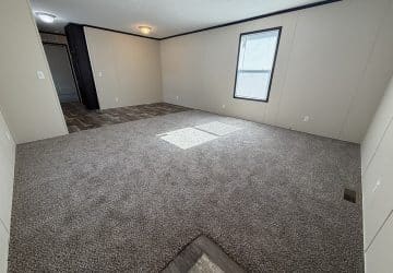 Empty room with light beige walls, a window letting in sunlight, gray carpet flooring, and an adjacent area with darker wood-like flooring leading to a doorway. The room has white outlets and minimal decor.