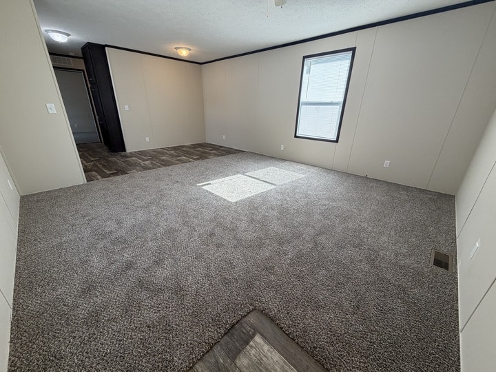 Empty room with light beige walls, a window letting in sunlight, gray carpet flooring, and an adjacent area with darker wood-like flooring leading to a doorway. The room has white outlets and minimal decor.
