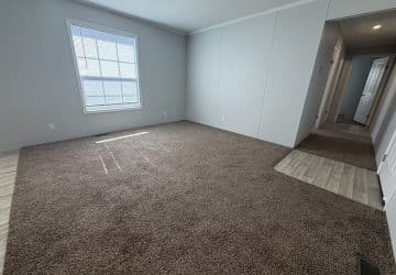 A bright, empty room with light gray walls, a large window, and brown carpet flooring. The carpet transitions to light wood flooring near a hallway that leads to other rooms.