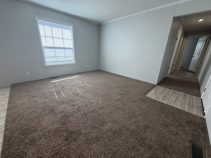 A bright, empty room with light gray walls, a large window, and brown carpet flooring. The carpet transitions to light wood flooring near a hallway that leads to other rooms.