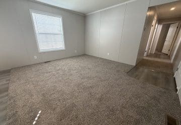 An empty room with light gray walls, a single window with closed blinds, carpeting, and a hallway with wood-look flooring leading to a white door.