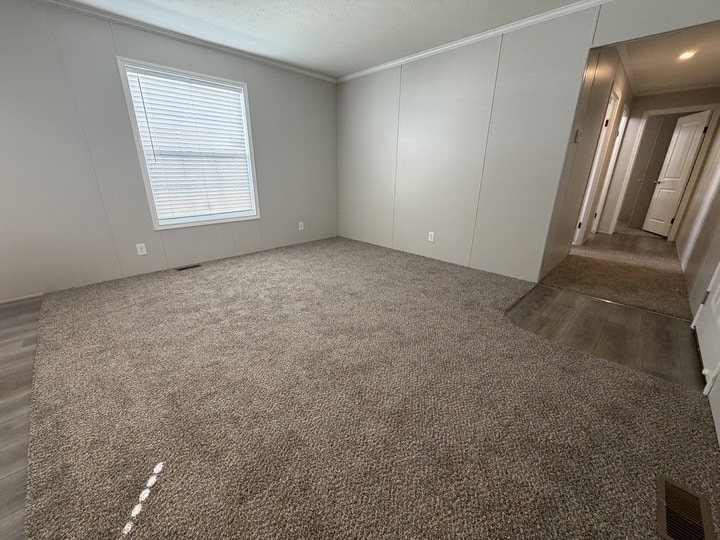 An empty room with light gray walls, a single window with closed blinds, carpeting, and a hallway with wood-look flooring leading to a white door.