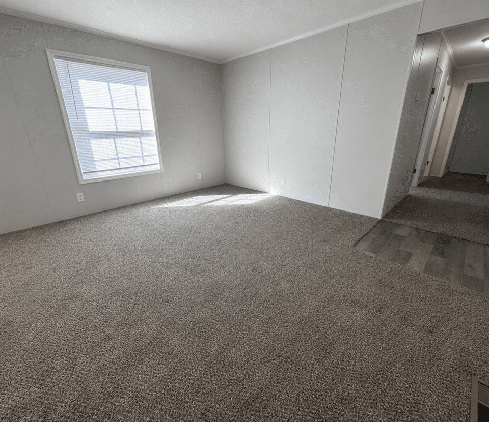 Empty room with light gray walls, a large window letting in sunlight, carpeted flooring, and a hallway with wood-style flooring leading to other rooms. The space appears clean and unfurnished.