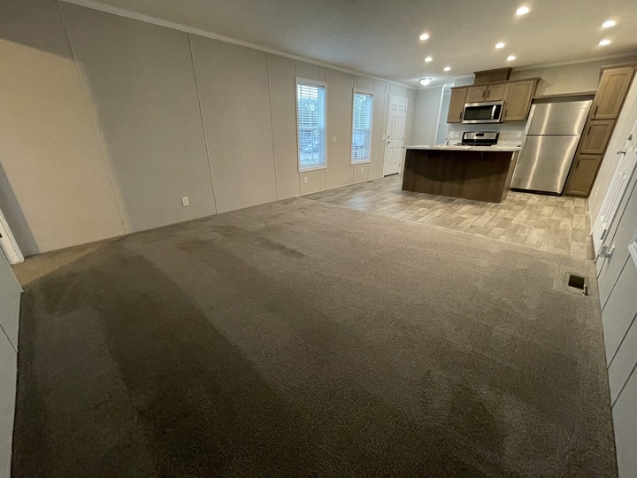 Open-plan living area with carpeted floor leading to a kitchen featuring wood cabinets, stainless steel appliances, and tile flooring. Two windows let in natural light; recessed ceiling lights provide illumination.