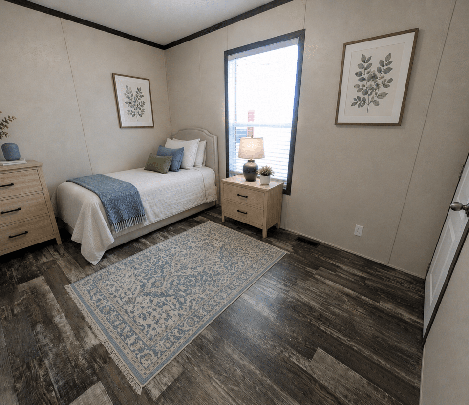 A neatly decorated bedroom with a single bed, light wood furniture, botanical art on the beige walls, a window letting in natural light, and a blue patterned rug on dark wood flooring.