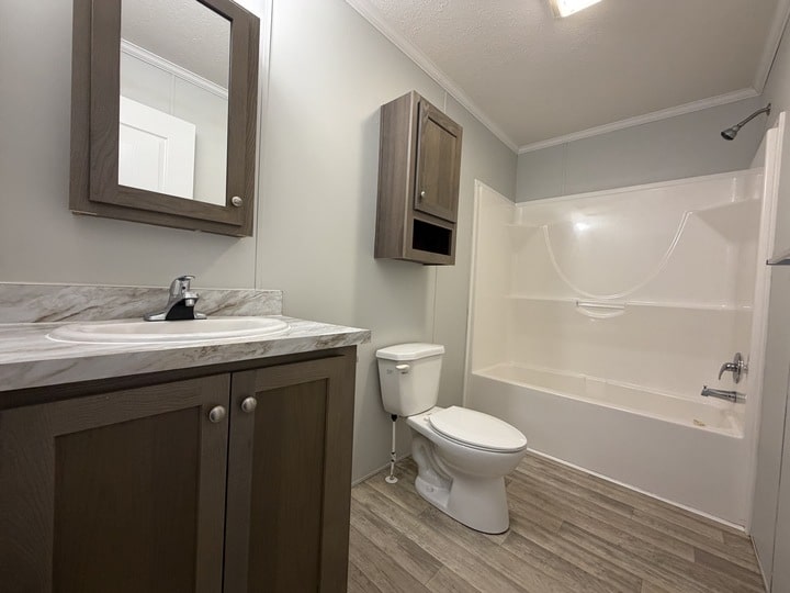 A modern bathroom with wood cabinets, a white sink, toilet, and a bathtub-shower combo. The floor has light wood-style tiles and the walls are light gray. A mirrored cabinet hangs above the sink.