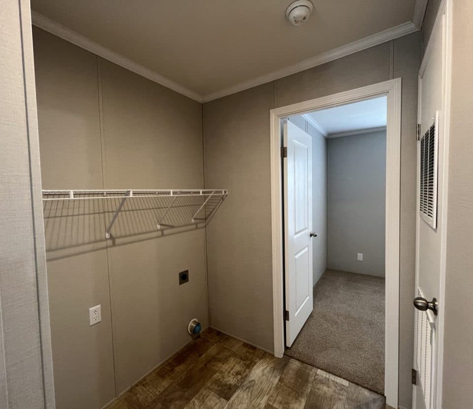 A small laundry room with beige walls, a wire shelf, utility hookups for a washer and dryer, and a door leading to a carpeted room. The floor is wood-patterned vinyl.