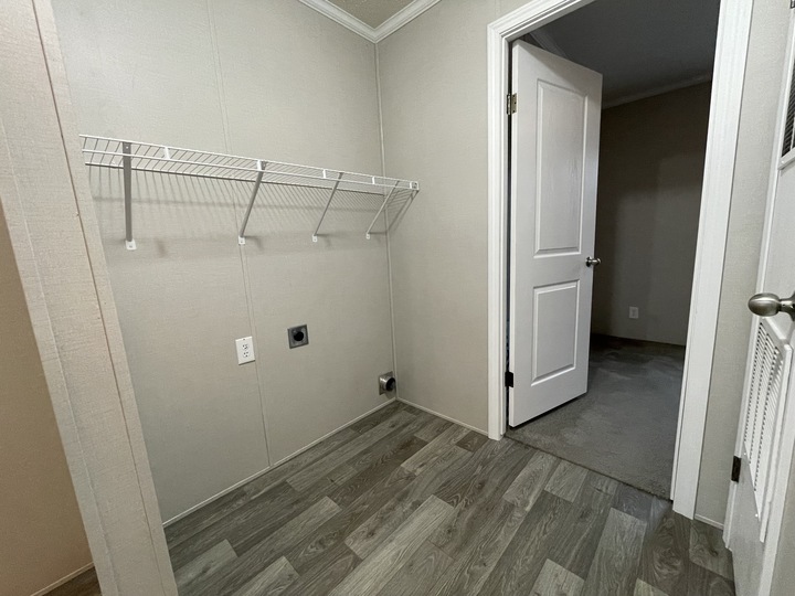 A small laundry room with light gray walls, a wire shelf, utility hookups on the wall, and wood-patterned flooring. An open door leads to a carpeted room.