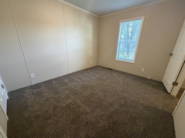 Empty room with beige walls, brown carpet flooring, a single window with blinds letting in natural light, and a white door partially open. The room appears clean and unfurnished.