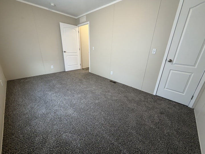 Empty room with beige walls, gray carpet, and two white doors—one open showing a lighted hallway, and one closed. The room has a simple, clean appearance with no furniture or decorations.