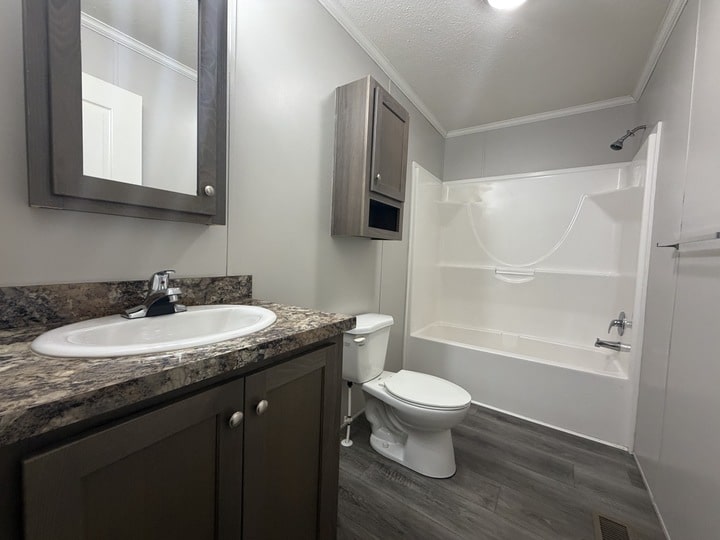 A modern bathroom with gray cabinets, a marble-patterned countertop with a sink, a toilet, wall-mounted storage, and a white bathtub-shower combo. The floor is gray wood-style, and the walls are light-colored.