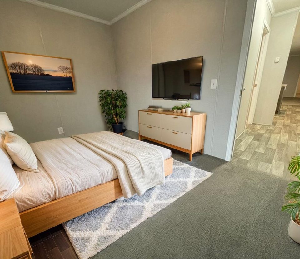 A modern bedroom with a wooden bed, white bedding, a dresser with a large TV, two green plants, a framed landscape photo on the gray wall, and an open hallway with wood-patterned flooring.