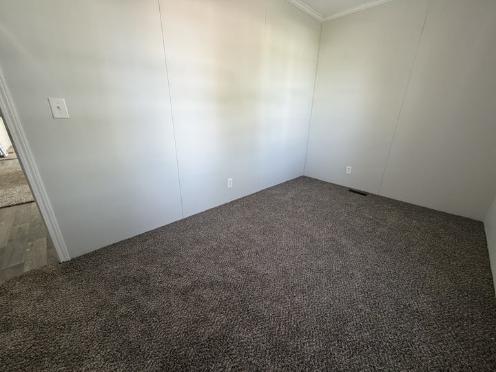 An empty room with light gray walls, white trim, and brown carpet flooring. There are electrical outlets on the walls and an open doorway visible on the left side.
