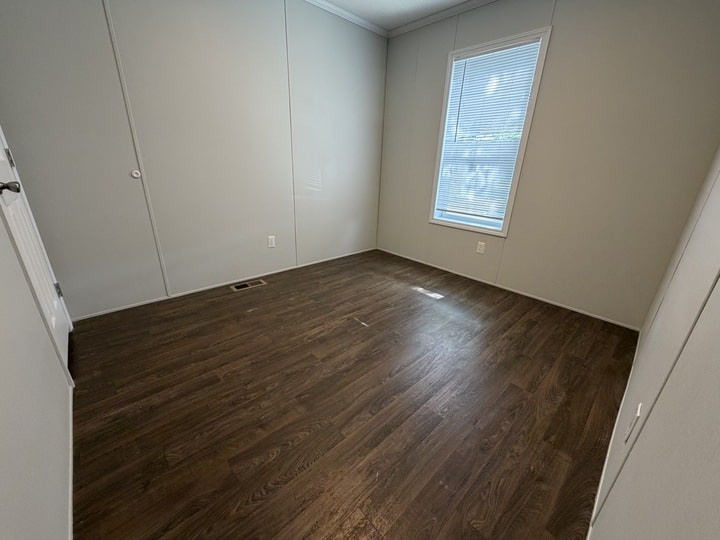 Empty room with light gray walls, dark wood flooring, a single window letting in natural light, and a white door slightly ajar on the left side.