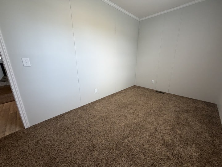 Empty room with light beige walls, white trim, beige carpet flooring, and two electrical outlets; a doorway opens to a space with light wood flooring on the left.