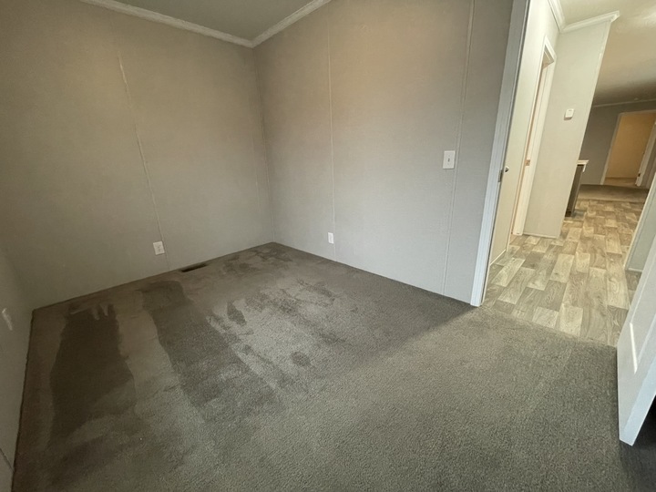 A small empty room with beige walls, gray carpet flooring, and a doorway leading to a hallway with wood-patterned tile. The carpet has visible clean and worn areas. The space appears well-lit and unfurnished.