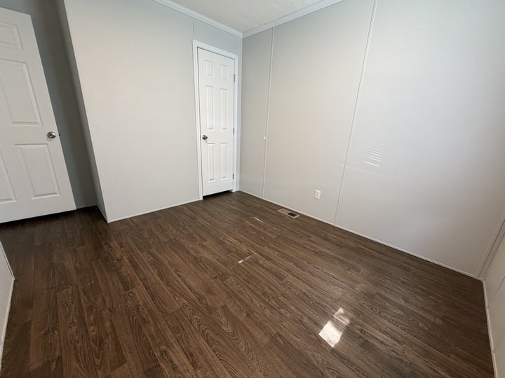 Empty room with light gray walls, dark wood flooring, two white doors, and a small floor vent. Sunlight is coming in from a window out of view on the right side.