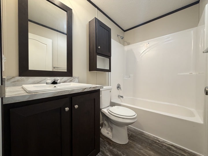 A modern bathroom with a dark wood vanity, a mounted mirror, a white sink, a toilet, and a white bathtub with a shower. The floor has wood-like tiles and the walls are light-colored.