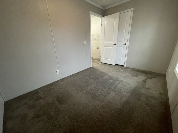Empty room with beige carpet and light-colored walls, featuring a double-door closet and an open doorway leading to another area with tile flooring and a laundry space in the background.