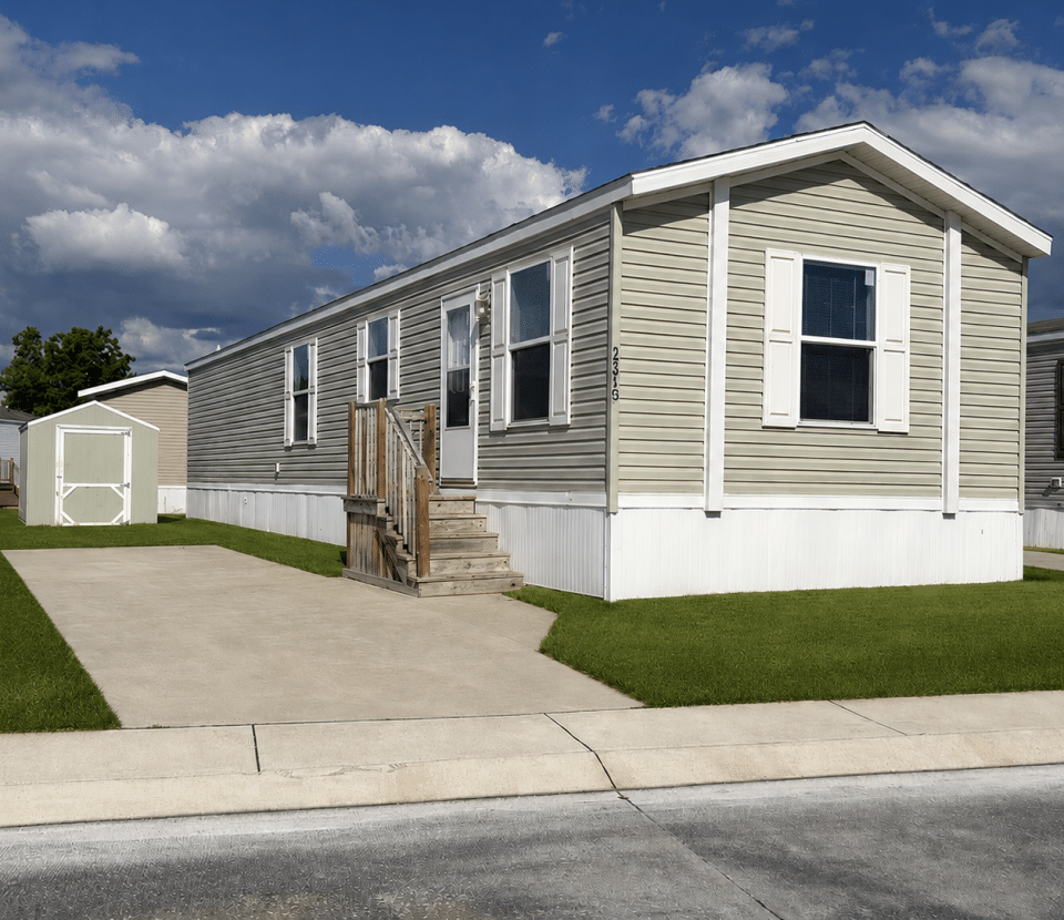 A beige mobile home with white trim and shutters sits on a neatly mowed lawn, with a small shed in the backyard and a concrete driveway in front under a partly cloudy sky.