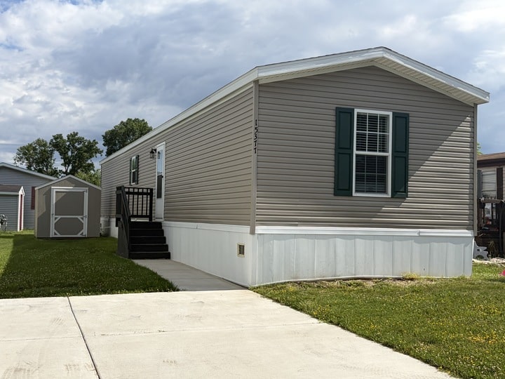 A beige mobile home with green shutters, a small porch with black steps, and a matching storage shed in the grassy yard under a partly cloudy sky. A concrete driveway is in the foreground.