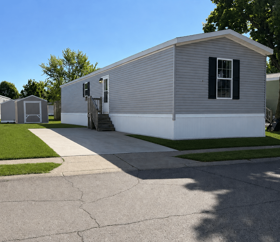 A gray manufactured home with white trim sits on a concrete driveway, with a small shed in the grassy yard and a tree nearby. The sky is clear and sunny.