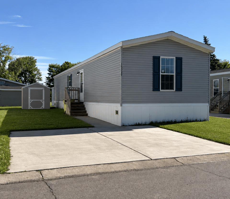 A light gray manufactured home with blue shutters sits on a grassy lot, with a concrete driveway in front and a small shed in the backyard under a clear blue sky.