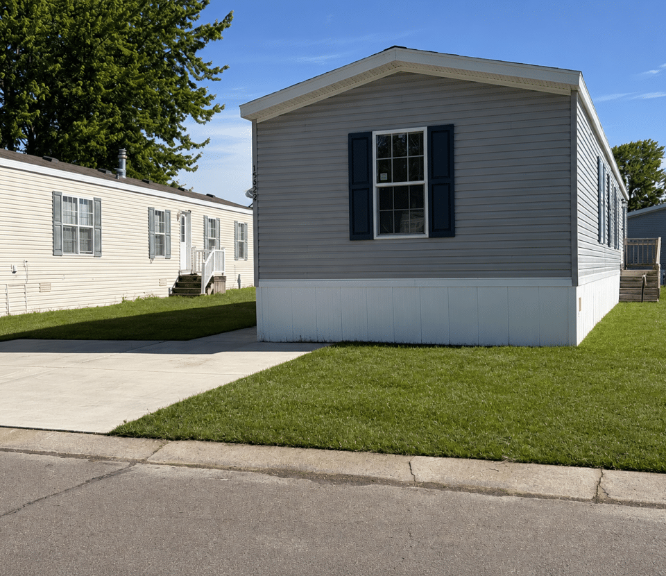 A gray mobile home with dark blue shutters sits on a green lawn next to a paved driveway, with another beige mobile home nearby and trees in the background under a clear blue sky.