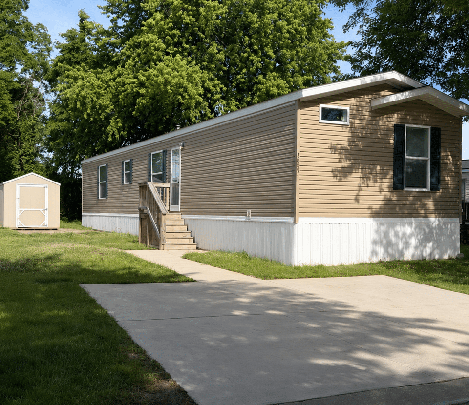 A beige mobile home with white trim and front steps sits on a concrete driveway, surrounded by green grass and trees. A small tan shed is visible in the background to the left.