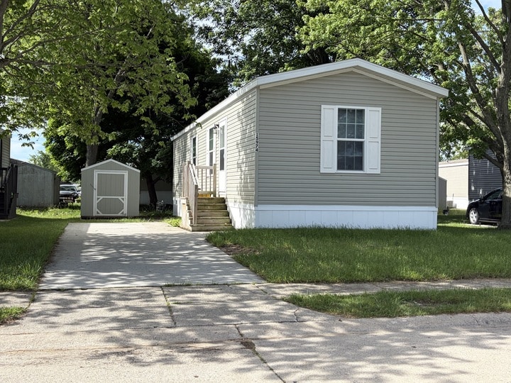 A light gray mobile home with white trim and shutters sits on a grassy lot with a concrete driveway leading to a small storage shed in the backyard, surrounded by trees and neighboring homes.