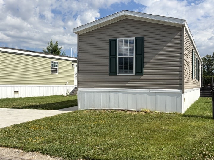 A beige mobile home with green shutters sits on a grassy lot with a driveway; another light green mobile home is visible in the background under a partly cloudy sky.