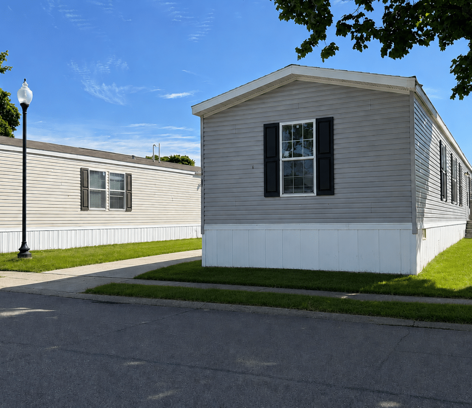 A light gray manufactured home with black shutters sits on a well-kept lawn next to a sidewalk under a blue sky, with another similar home and a streetlamp nearby.