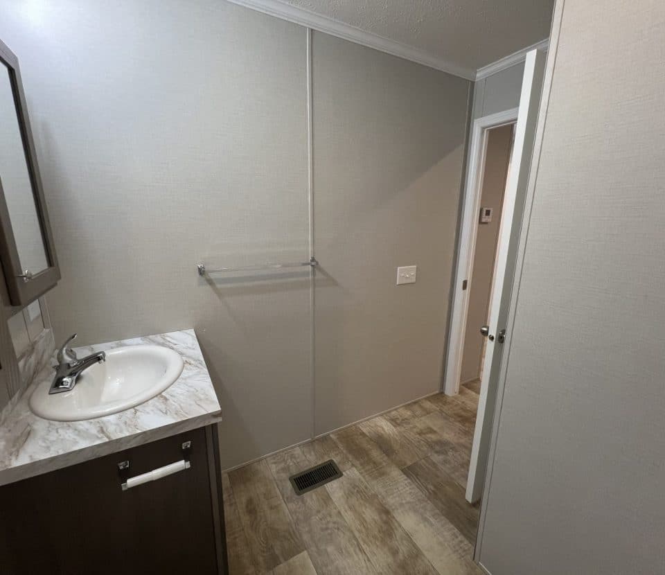 A bathroom with a marble countertop sink, a wall-mounted cabinet, a towel rack, and light wood flooring. The walls are light gray, and there is a white door leading to another room.