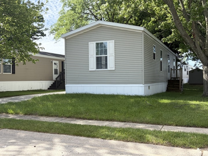 A beige mobile home with white shutters and skirting sits on a green lawn next to a sidewalk, shaded by large trees. Another similar home is visible to the left.