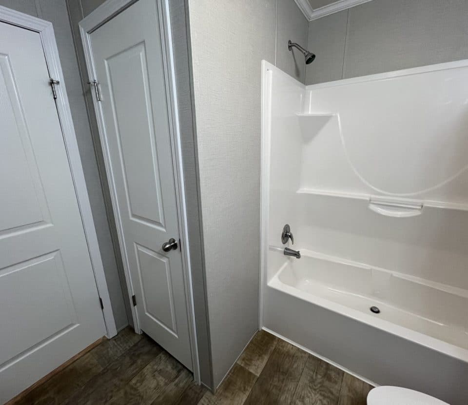 A small bathroom with two white doors, a white bathtub and shower combination with a built-in shelf, and wood-look flooring.
