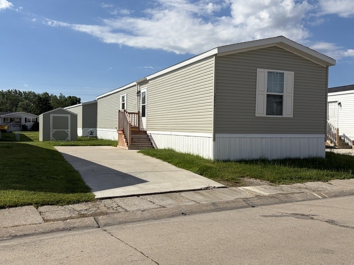 A beige manufactured home with white trim and a small front porch sits on a concrete driveway next to a grassy lawn, under a blue sky with scattered clouds.