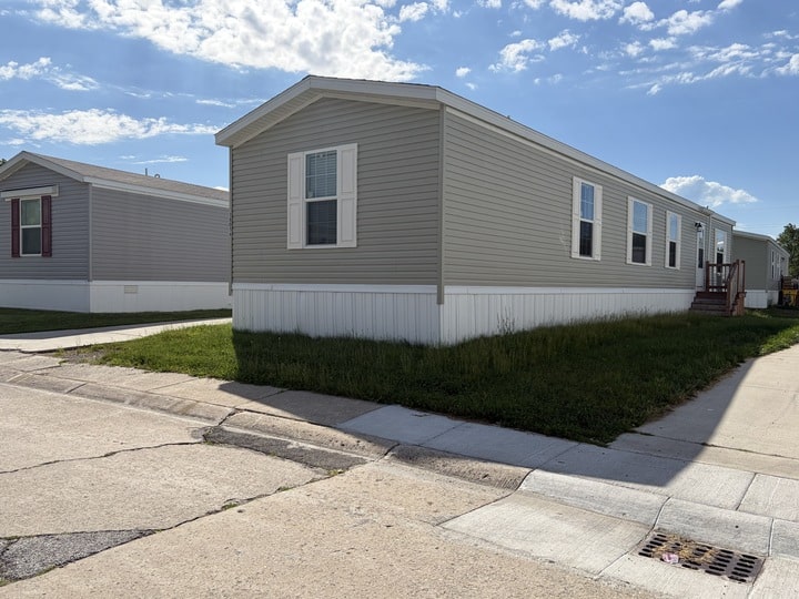 A light gray manufactured home with white trim sits on a grassy lot near a sidewalk under a partly cloudy sky. Another similar home is visible in the background.