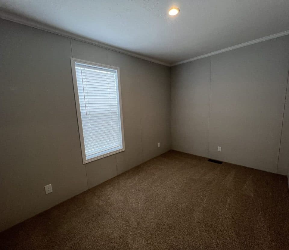 A small, empty room with beige walls, brown carpet flooring, a single window with closed blinds, and a ceiling light turned on. The room appears clean and simple with minimal decoration.